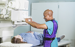 Older bald black man setting up a machine to x-ray a white woman with brown hair laying on a table